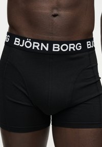 Μαύρα ανδρικά μποξεράκια με λευκή ζώνη μέσης με το λογότυπο "BJÖRN BORG", εφαρμοστά σε σκούρο ανδρικό κορμό από τη μέση έως τη μέση του μηρού.