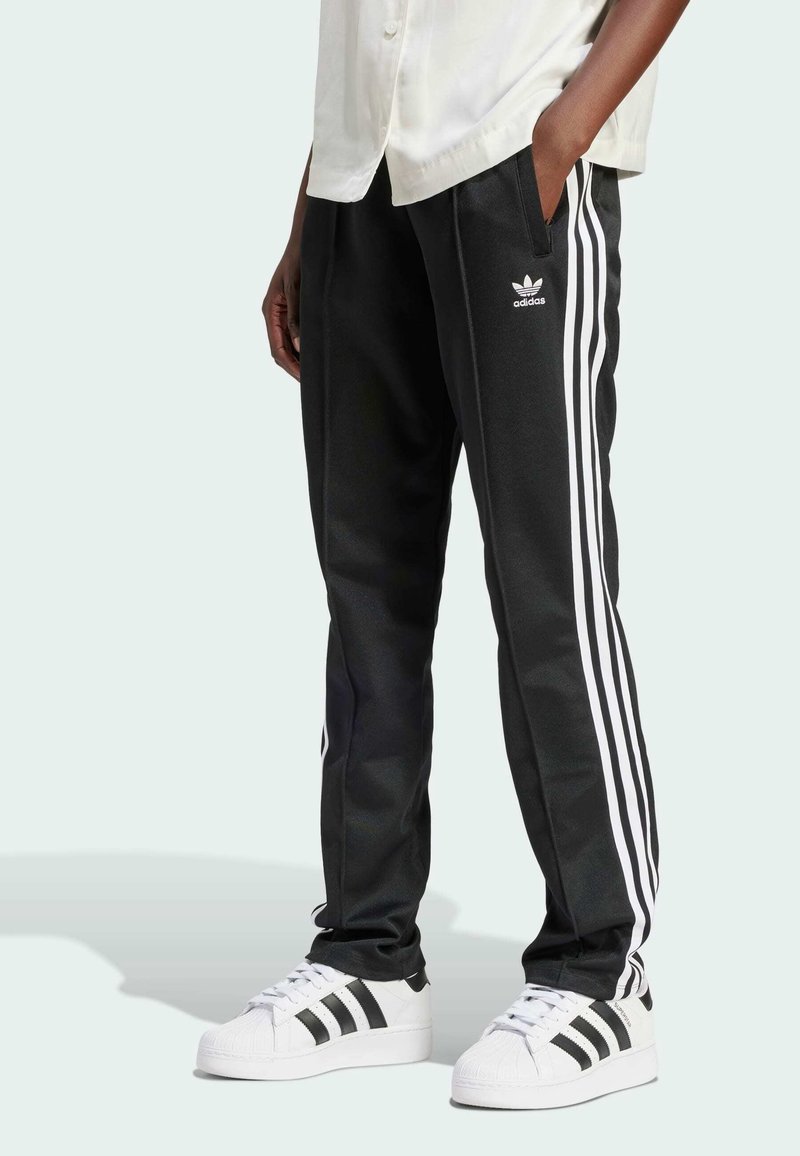 Czarne spodnie sportowe z trzema białymi paskami po każdej stronie, wykonane z gładkiego materiału, w połączeniu z białymi butami sportowymi z czarnymi paskami.