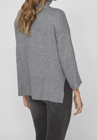 Grau gestrickter Pullover mit Rollkragen, mit geripptem Muster und seitlichen Schlitzen. Weiches Material mit lockerem Schnitt, kombiniert mit dunklen Jeans.