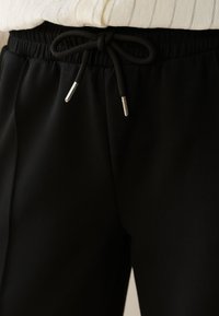 Gros plan sur un pantalon taille élastique noir avec un cordon noir noué doté d'embouts métalliques, associé à un haut rayé de couleur claire.