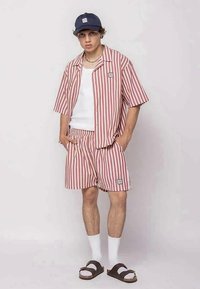 Young man standing with hands in pockets wearing red and white vertical striped shirt and shorts, white tank top, navy cap, white socks, and sandals.