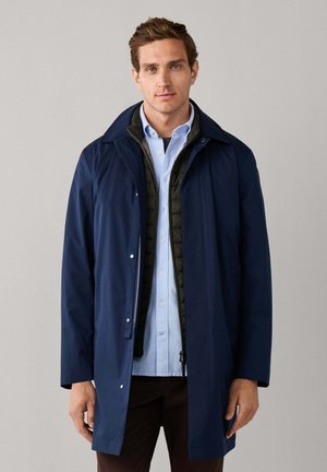 Hombre con impermeable azul marino en capas, chaleco negro acolchado y camisa azul claro abotonada sobre un fondo liso.