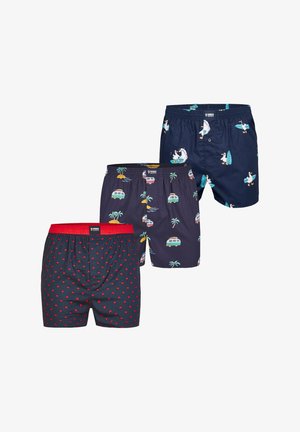 Trois paires de boxers : une avec des cœurs rouges sur un fond sombre, une autre avec des palmiers et des vans vintage, et une unie marine avec des motifs de surf.