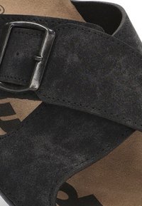 Sandalia de ante negra con una tira ancha y un hebilla de metal plateado, plantilla suave y suela tostada con logo. Superficie texturizada evidente.