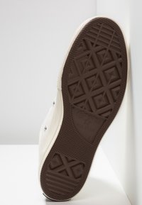 Canvas-sneaker med en beige overdel, brun gummisåle og diamantmønster. Har metalløyler og "Converse All Star" merke.
