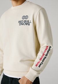 Crème-kleurige sweatshirt met ribgebreide mouwen en onderrand. Bevat bedrukte graphics in zwart en rood: "RED BULL RACING" en "Authentic Race Gear".
