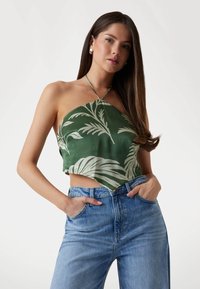 Vihreä crop-top, jossa on valkoisia lehtikuvioita ja ohuet kaula-aukkosangat, yhdistettynä vaaleansinisiin korkeavyötäröisiin farkkuihin. Sujuva tekstuuri.