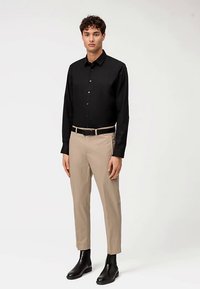 Zwart shirt met lange mouwen en knopen, beige op maat gemaakte broek en zwarte enkellaarzen, staand tegen een effen achtergrond.