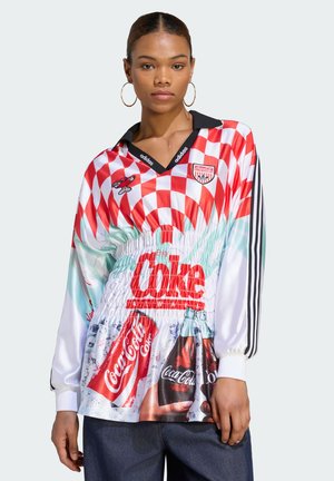 Ung kvinde iført langærmet Adidas-trøje med rød-hvid ternet top og Coca-Cola flaske og dåse grafik på fronten.