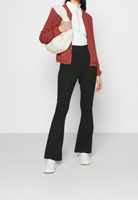 Veste bomber rouge à fermeture éclair portée sur un haut blanc à manches longues, associée à un pantalon noir évasé et des baskets blanches, avec un sac à bandoulière crème à texture.