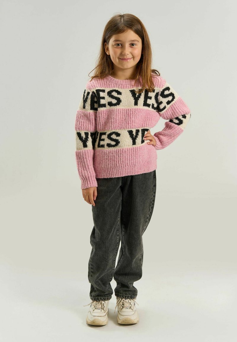 Jeune fille aux cheveux bruns, debout en souriant, portant un pull rayé rose et blanc avec le texte « YES », pantalon noir et baskets blanches.
