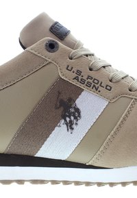Beige urheilukengät, joissa on sileä synteettinen päällinen, verkkokankaisia yksityiskohtia, logo ja valkoinen raita. Kumipohja, jossa on teksturoitu pito.
