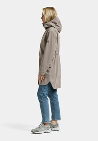 Vaaleanharmaa vedenpitävä takki hupulla, kapealla vyötäröllä ja napeilla koristeltu. Päällä sinisten farkkujen ja harmaiden tennareiden kanssa. Sivukuva.