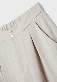 Beżowe lniane spodnie z elastycznym paskiem w talii, dwoma białymi guzikami z przodu, boczną kieszenią oraz plisą z przodu.