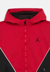 Κόκκινο και μαύρο αδιάβροχο μπουφάν με φερμουάρ μπροστά, κουκούλα και επένδυση από δίχτυ. Διαθέτει το μαύρο λογότυπο Jumpman και λευκή διαγώνια ραφή.