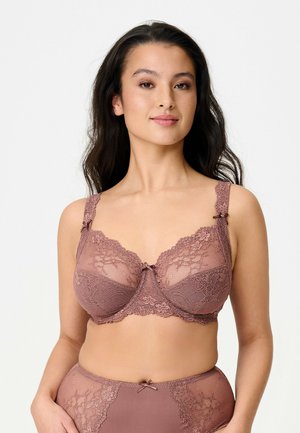 Kvinde med langt mørkt hår iført mauve blondetop og matchende højtaljede blondeunderbukser, stående mod en ensartet lys baggrund.
