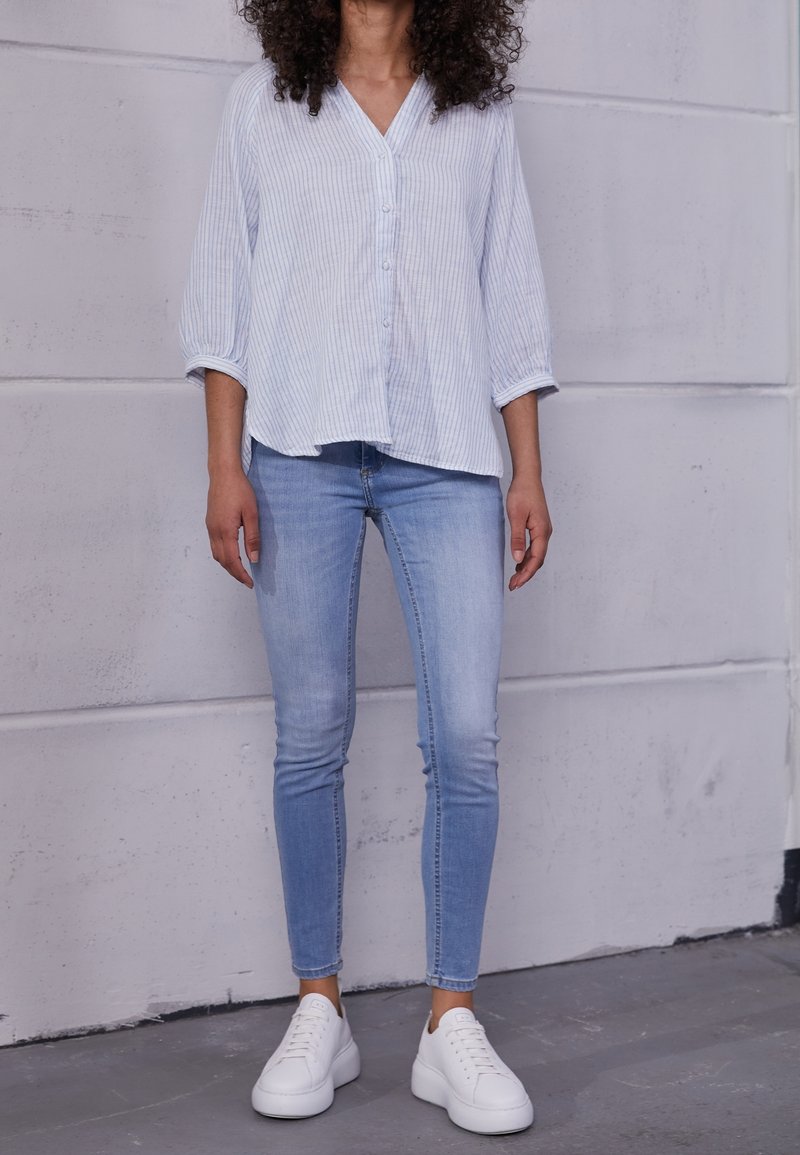 Jasnoniebieska koszula na guziki w paski z dekoltem w serek, zestawiona z jasnoniebieskimi obcisłymi dżinsami i białymi tenisówkami. Gładka tkanina i casualowy krój.
