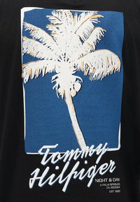 Λευκό φοινικόδεντρο με καρύδες σε μπλε ορθογώνιο φόντο, με το κείμενο "Tommy Hilfiger" και πρόσθετη μικρή εκτύπωση από κάτω.