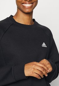 Persona sonriente con un sudadera negra de Adidas, manos suavemente entrelazadas cerca del pecho, logo visible en el lado izquierdo.