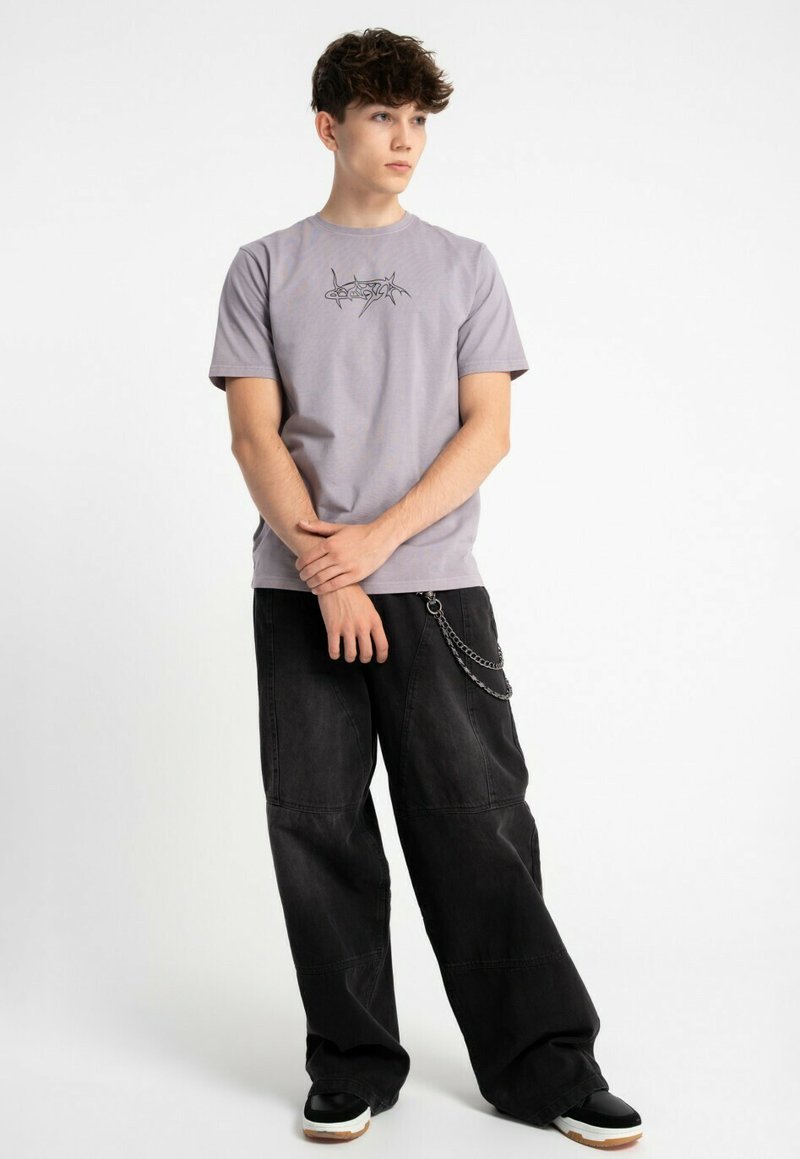 Jasnoszara bawełniana koszulka z czarnym nadrukiem na piersi, zestawiona z luźnymi czarnymi spodniami z detalem łańcucha i kontrastowymi wykończeniami.