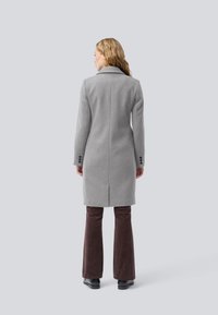 Palton gri din lână cu o croială ajustată, mâneci lungi și o deschidere pe spate. Asortat cu pantaloni flared din bumbac cu țesătură în carouri de culoare maro și pantofi negri.