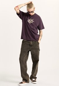 Purple t-shirt with a star-shaped graphic, paired with loose-fitting, faded brown jeans and two-tone sneakers. Simple sunglasses worn.