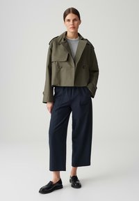 Geknipt olijfgroen jack met dubbelgeknoopte knopen, grijs shirt eronder, gecombineerd met marineblauwe wijde broek en zwarte loafers.