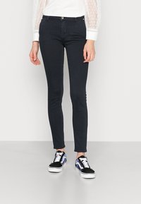 Jeans slim fit em azul-marinho, feitos de um tecido suave e elástico, combinados com sapatilhas pretas e azuis com padrão, com solas e atacadores brancos.