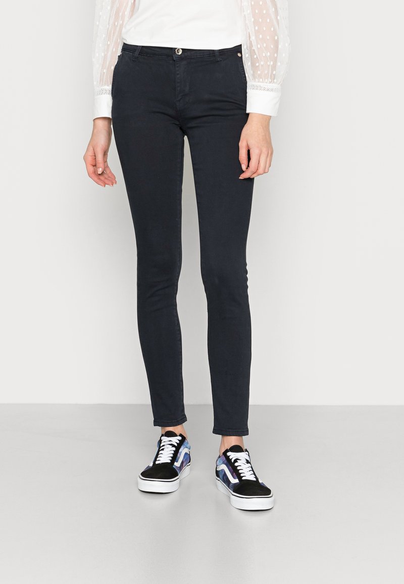 Jeans slim fit em azul-marinho, feitos de um tecido suave e elástico, combinados com sapatilhas pretas e azuis com padrão, com solas e atacadores brancos.