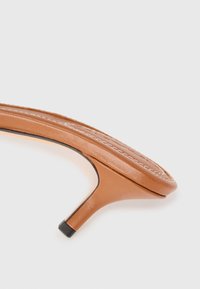 Tan high-heeled sandal with a smooth leather upper, curved design, and a black accent on the heel. Stitch detailing around the edge.