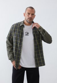 Plaid shirt in green and navy with gray long sleeves, over a white T-shirt featuring an embroidered nautical design. Black pants.