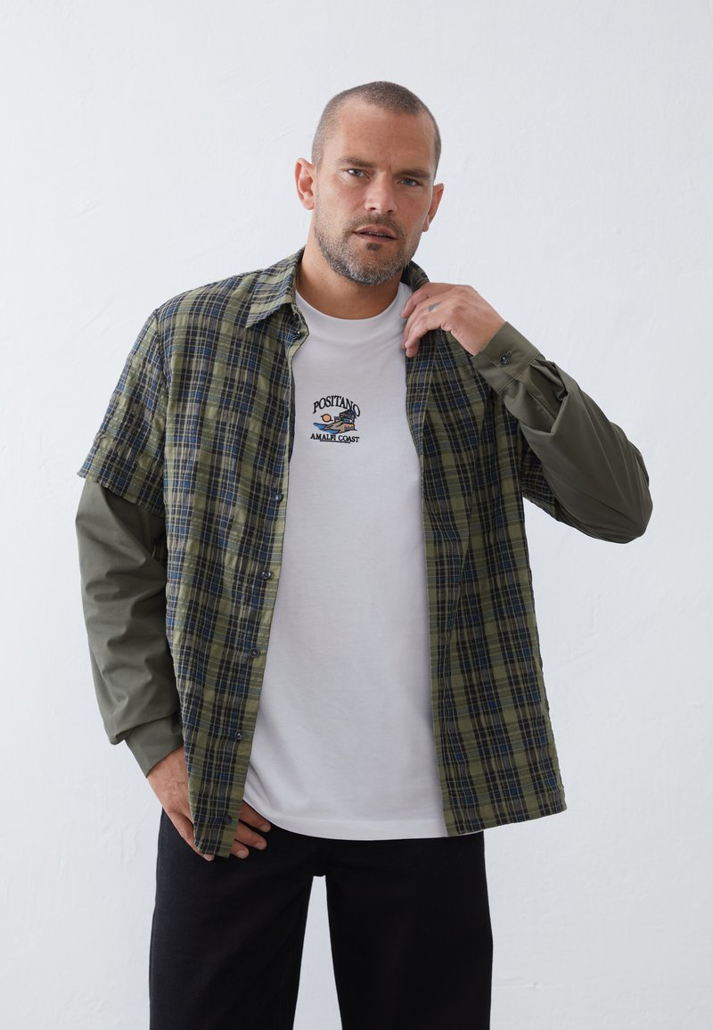 Plaid shirt in green and navy with gray long sleeves, over a white T-shirt featuring an embroidered nautical design. Black pants.