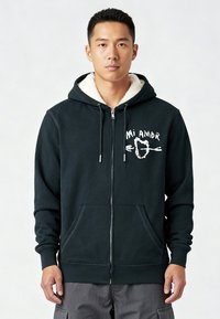 Donkergroene rits-hoodie met een zachte crèmekleurige voering, voorzakken en een witte tekstgrafiek met de tekst "Mi Amor." Zacht materiaal met geribbelde manchetten.