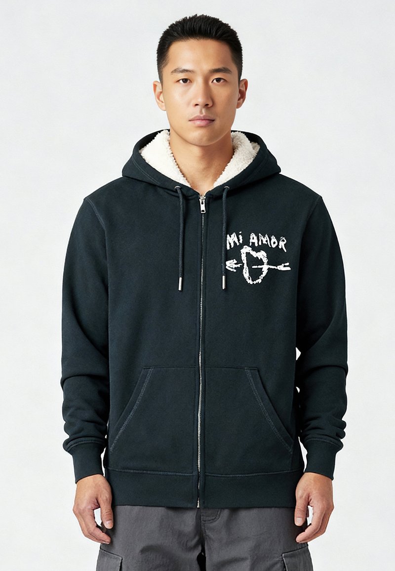 Donkergroene rits-hoodie met een zachte crèmekleurige voering, voorzakken en een witte tekstgrafiek met de tekst "Mi Amor." Zacht materiaal met geribbelde manchetten.