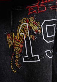Tigre ricamato con strisce rosse e gialle che si arrampica su una tasca di denim nero con il numero "19" contornato in bianco.