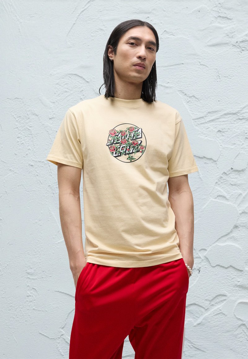 Beige cotton t-shirt featuring a circular graphic with red roses and green leaves. Paired with red trousers, showcasing a relaxed fit.