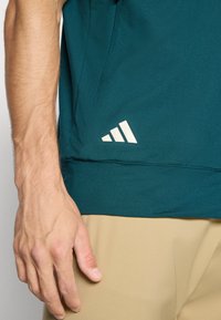 Male hand resting beside dark teal shirt with white logo above beige pants, showing casual clothing and relaxed posture.