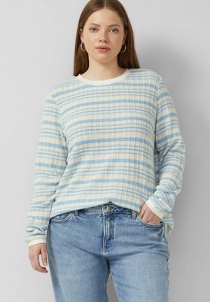 Vrouw met steil lang haar die zilveren oorringen draagt, een blauw-wit gestreepte trui en lichtblauwe jeans, poserend met één hand in de zak.