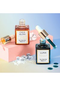 Deux flacons en verre d'huiles pour le visage sur un bloc rose. L'un est en ambre avec un compte-gouttes ; l'autre est bleu avec un compte-gouttes bleu clair. Des cristaux transparents les entourent.