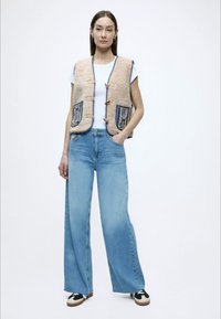 Μπεζ γιλέκο με καπιτονέ ύφασμα και πολύχρωμες τσέπες με σχέδια, φορεμένο πάνω από λευκό τοπ και συνδυασμένο με ανοιχτό μπλε παντελόνι φαρδιάς γραμμής και μαύρα αθλητικά παπούτσια.