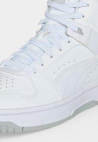 Κοντινή λήψη ενός λευκού sneaker ψηλού τύπου με υφή δέρματος, οπές στο μπροστινό μέρος και παχιές λευκές κορδόνια δεμένα πάνω από τη γλώσσα.
