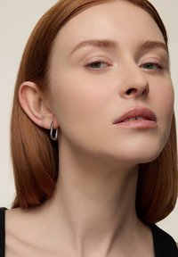 Srebrne kolczyki koła o smukłym, zakrzywionym kształcie z teksturowanymi detalami, prezentowane na modelce z półdługimi prostymi włosami.
