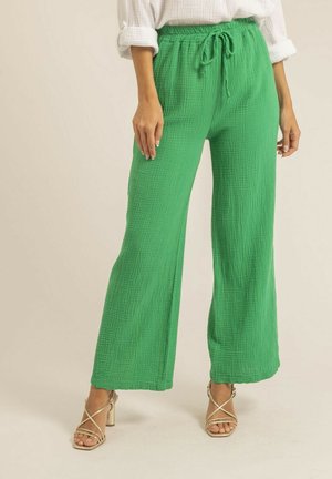 Femme portant un pantalon ample et texturé de couleur vert vif avec une taille à cordon de serrage et des sandales à talons hauts à lanières beige, se tenant devant un fond uni.