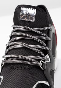 Czarne trampki z białymi i czarno-szachownicowymi sznurówkami, srebrnym logo "MK" na języku oraz białymi i czerwonymi akcentami na podeszwie.