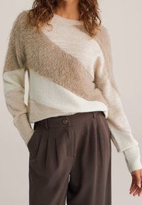 Pull rayé beige clair et crème avec des accents diagonaux duveteux, en tissu tricoté texturé, et des poignets côtelés, associé à un pantalon plissé marron foncé.