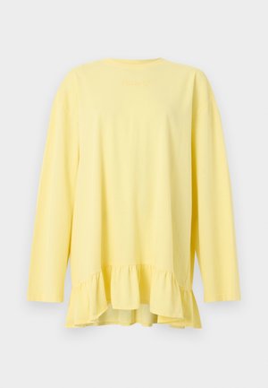 Vrouw die een losse gele jurk met gerufflede zoom draagt, een zwarte minirok, zwarte laarzen en een zwarte baseballpet, staand tegen een witte achtergrond.