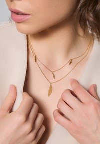 Złocona naszyjnik składający się z trzech łańcuszków z małymi zawieszkami w kształcie piór. Modelka ma na sobie jasną bluzkę, ręce spoczywają przy dekolcie.