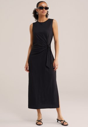 Žena v čiernych midi šatách bez rukávov s bočným uzlom, čiernych sandáloch, kruhových náušniciach a obdĺžnikových slnečných okuliaroch na jednofarebnom pozadí.