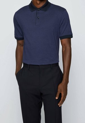 Tummansininen polo-paita, jossa on teksturoitu kangas, musta kaulus ja lyhyet hihat. Yhdistetty räätälöityihin mustiin housuihin, kädet taskuissa.