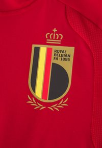 Tecido vermelho com um emblema dourado featuring uma coroa, listras verticais pretas, amarelas e vermelhas, texto "FA REAL BELGA 1895" e folhas de louro.